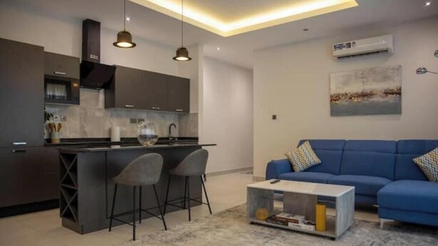 A modern kitchen with dark cabinets, a central island, and bar stools. There is a blue sofa with patterned cushions in the living area.