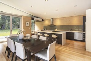 A modern kitchen with a central island, a dining area with a dark wood table and white chairs, and a view of a garden through large windows.