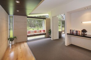 A modern interior with a wooden floor, a bench with cushions, a bookshelf, and a view of greenery outside.
