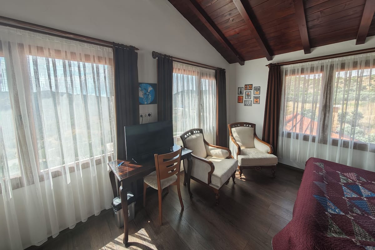 Una habitación con techo de madera, un escritorio con computadora, dos sillones tapizados, un sofá rojo y ventanas amplias con cortinas translúcidas.