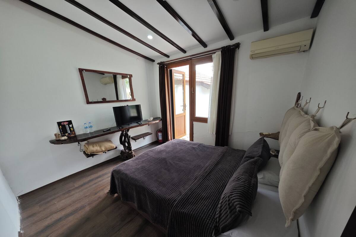 Un dormitorio con cama, televisor, estante con objetos, espejo y ventana.