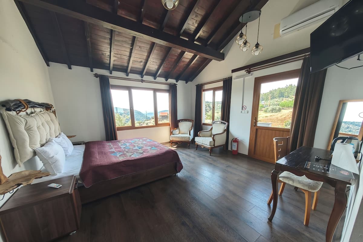 Un dormitorio con techo de madera, una cama con colcha, un escritorio, dos sillas y vistas al exterior.