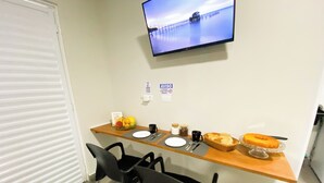 Een hotelkamer met een eethoek, met een houten tafel, zwarte stoelen, ontbijtartikelen en een aan de muur bevestigde televisie.
