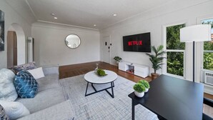 Ein modernes Wohnzimmer mit einer Couch, einem runden Couchtisch, einem an der Wand montierten Fernseher und einem großen Fenster mit Blick auf Grünflächen.