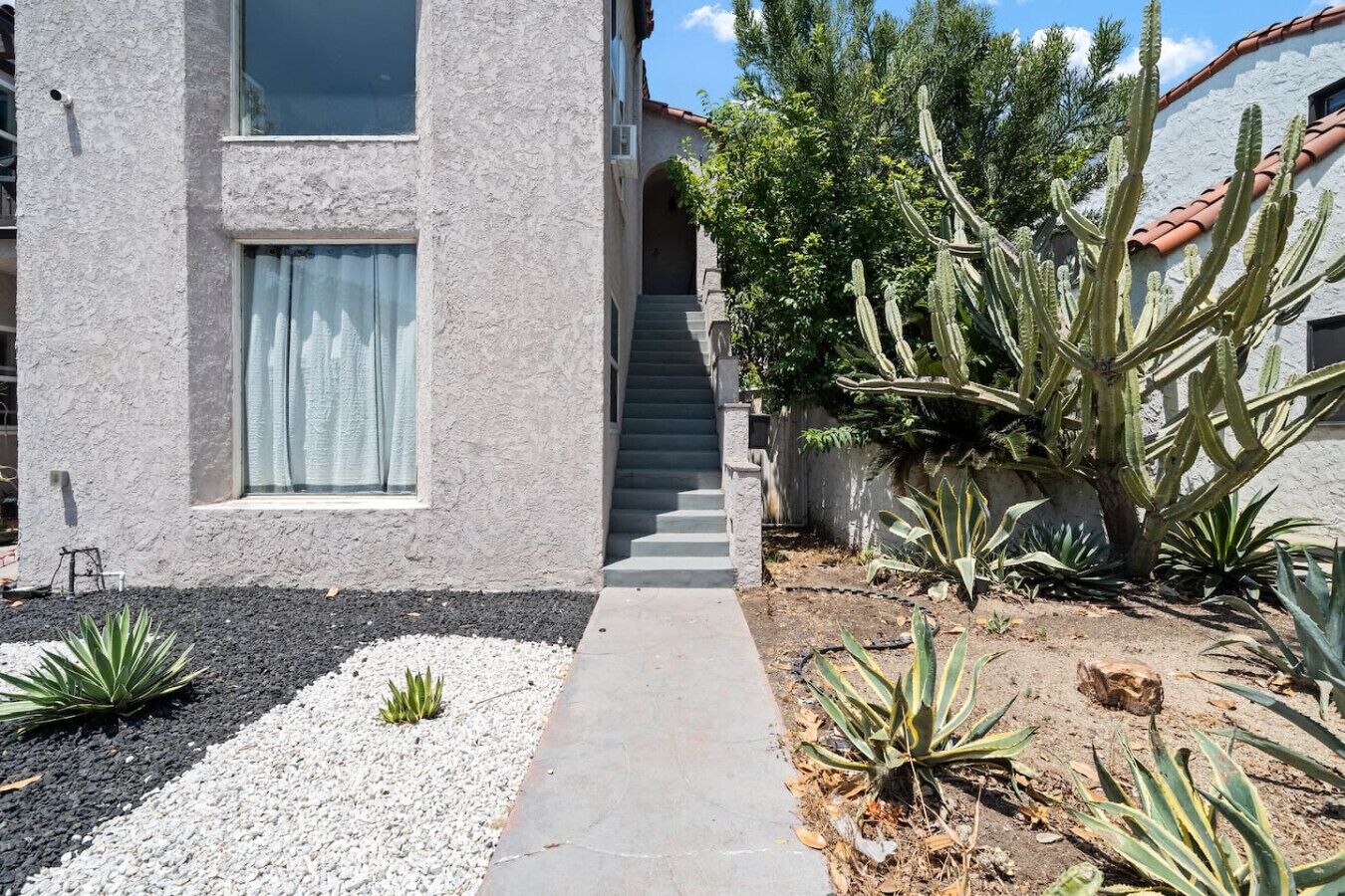 Een gebouw met een betonnen gevel, een trap die naar een ingang leidt, en een tuin met cactussen en agaveplanten.