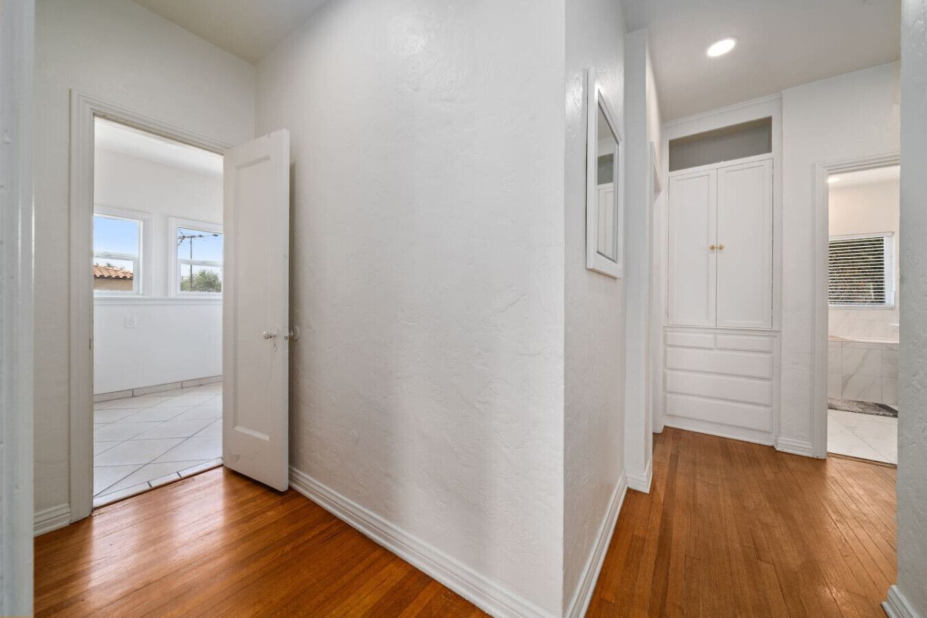 A hallway with white walls and wooden flooring, leading to a door and a room with a large mirror.