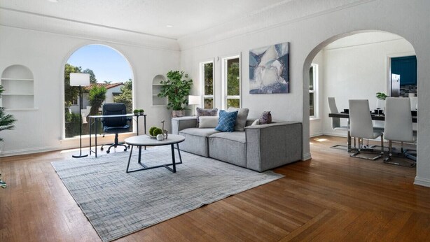 Ein geräumiges Wohnzimmer mit grauem Sofa, einem Couchtisch und einem Essbereich mit Stühlen. Große Fenster bieten einen Blick ins Freie.