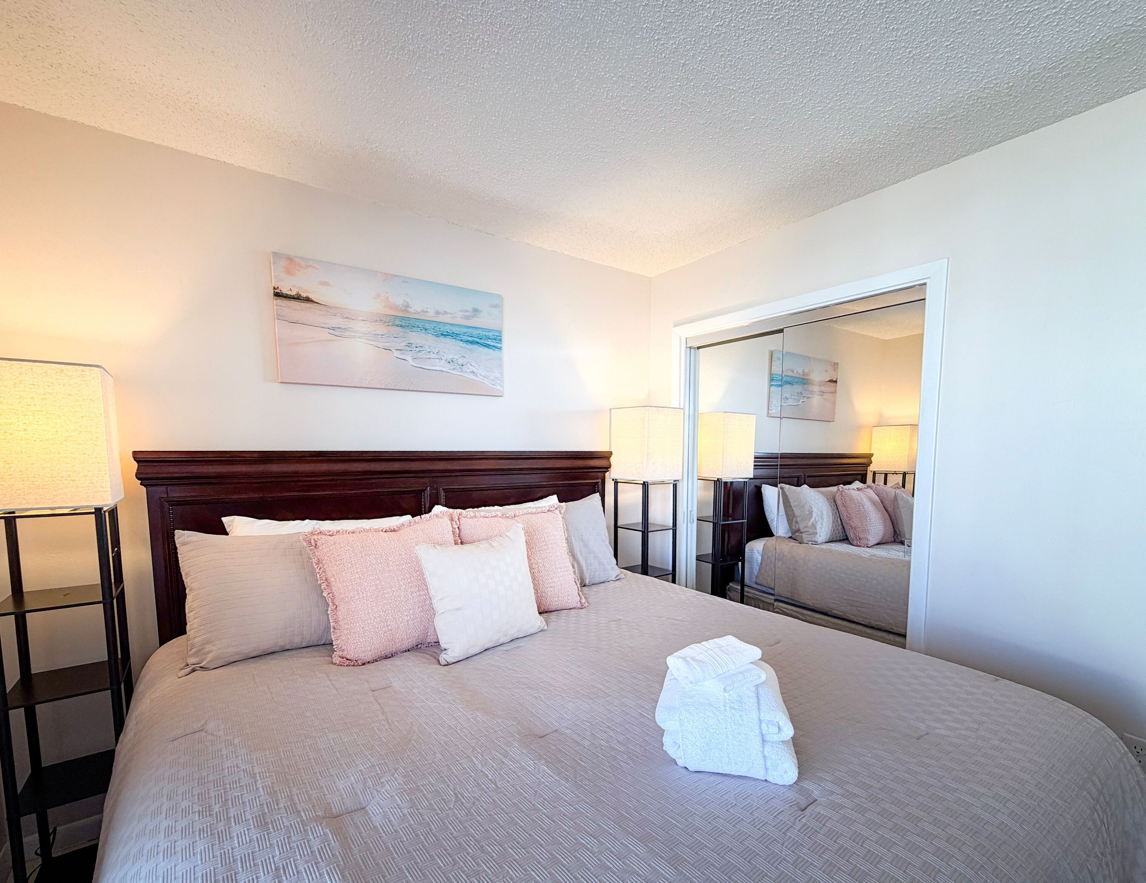 A hotel room with a large bed, two bedside tables with lamps, a headboard, a painting on the wall, and a mirror.