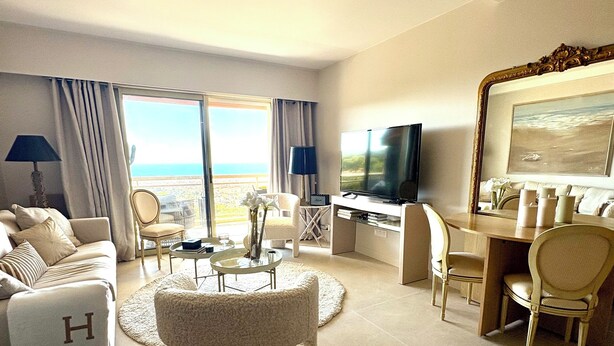 Une chambre d ’hôtel spacieuse avec un grand lit, un coin salon avec un téléviseur, une table à manger et un balcon offrant une vue sur la mer.