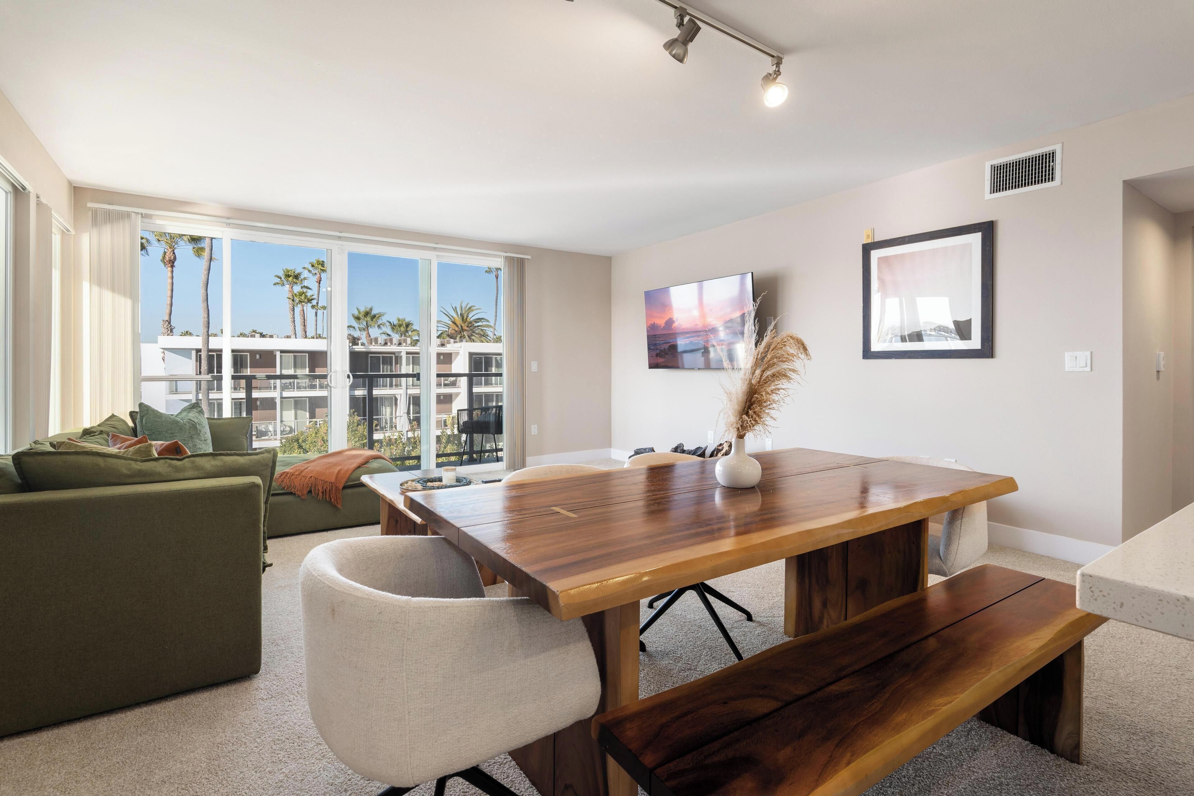 A modern living room with a wooden dining table, a green sofa, and a view of palm trees through large windows.