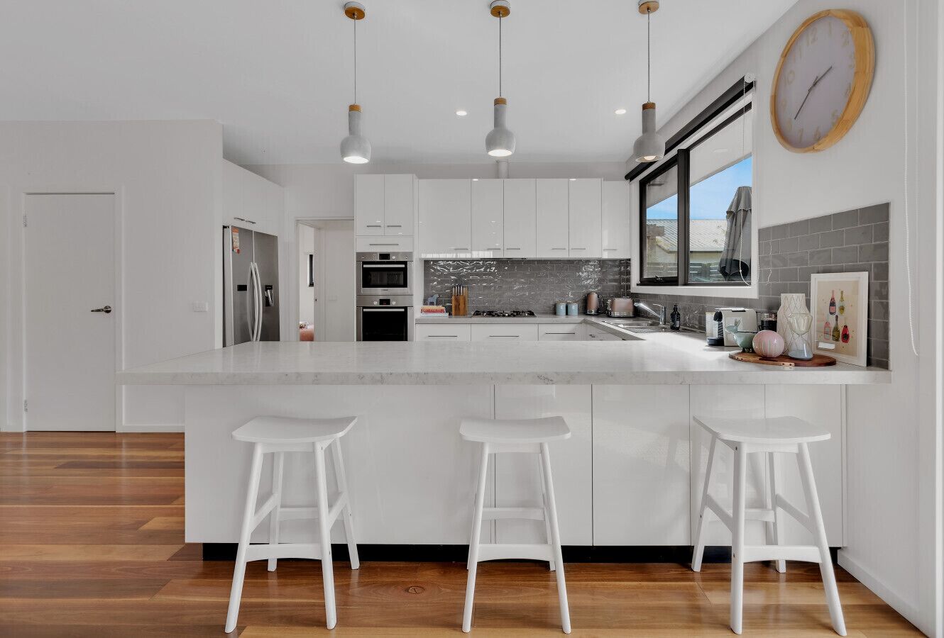 A modern kitchen with white cabinetry, a large island with stools, pendant lighting, and a view through the window.