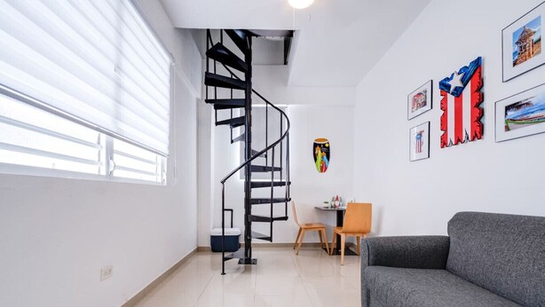 Un salon moderne avec un canapé gris, une table à manger en bois accompagnée de deux chaises, un escalier en colimaçon noir et un mur orné de tableaux.