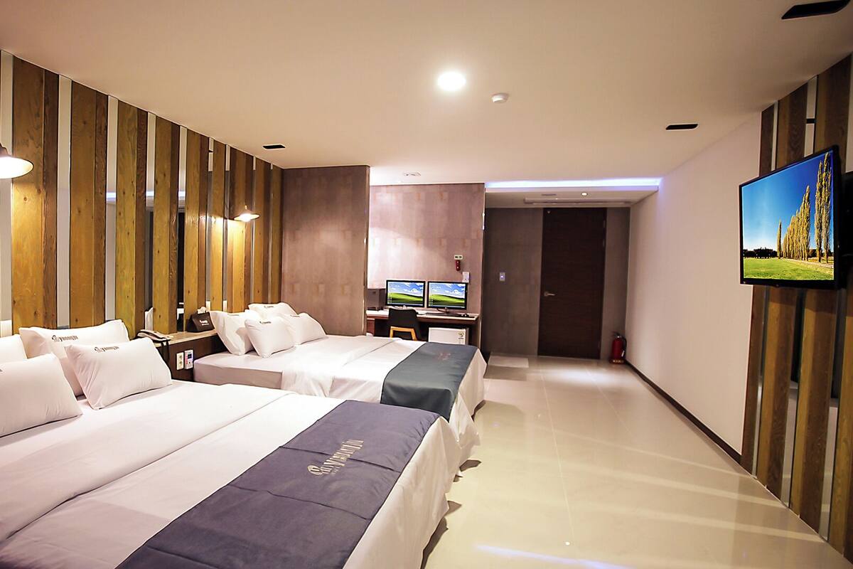 A hotel room with two beds, a desk with a computer, a TV, and a wooden wall paneling.