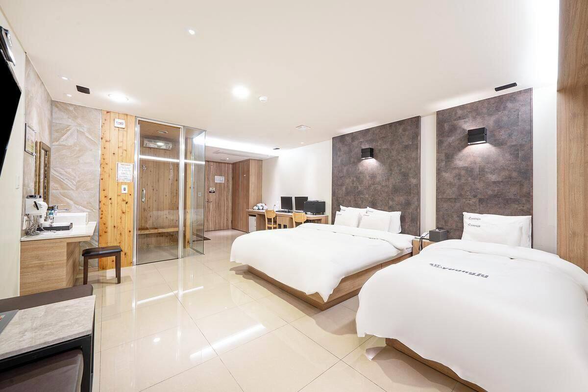 A modern hotel room with two beds, a desk, a chair, and a bathroom with a mirror and sink.