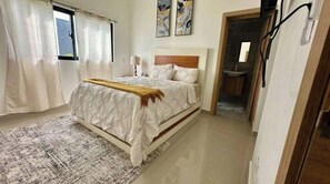 Quarto com cama, mesa de cabeceira, janela com cortinas e banheiro visível através de uma porta aberta.