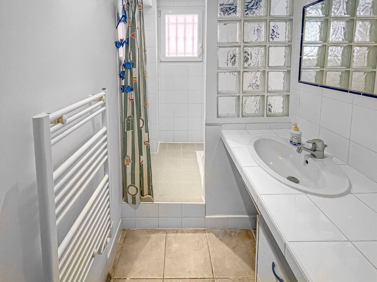 Een badkamer met een witte radiator, een douchegordijn met een patroon, een wastafel met een kraan en een betegelde muur met een raam.