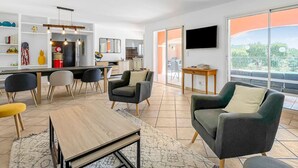 A modern living room with a dining area, large windows, and a kitchen in the background.