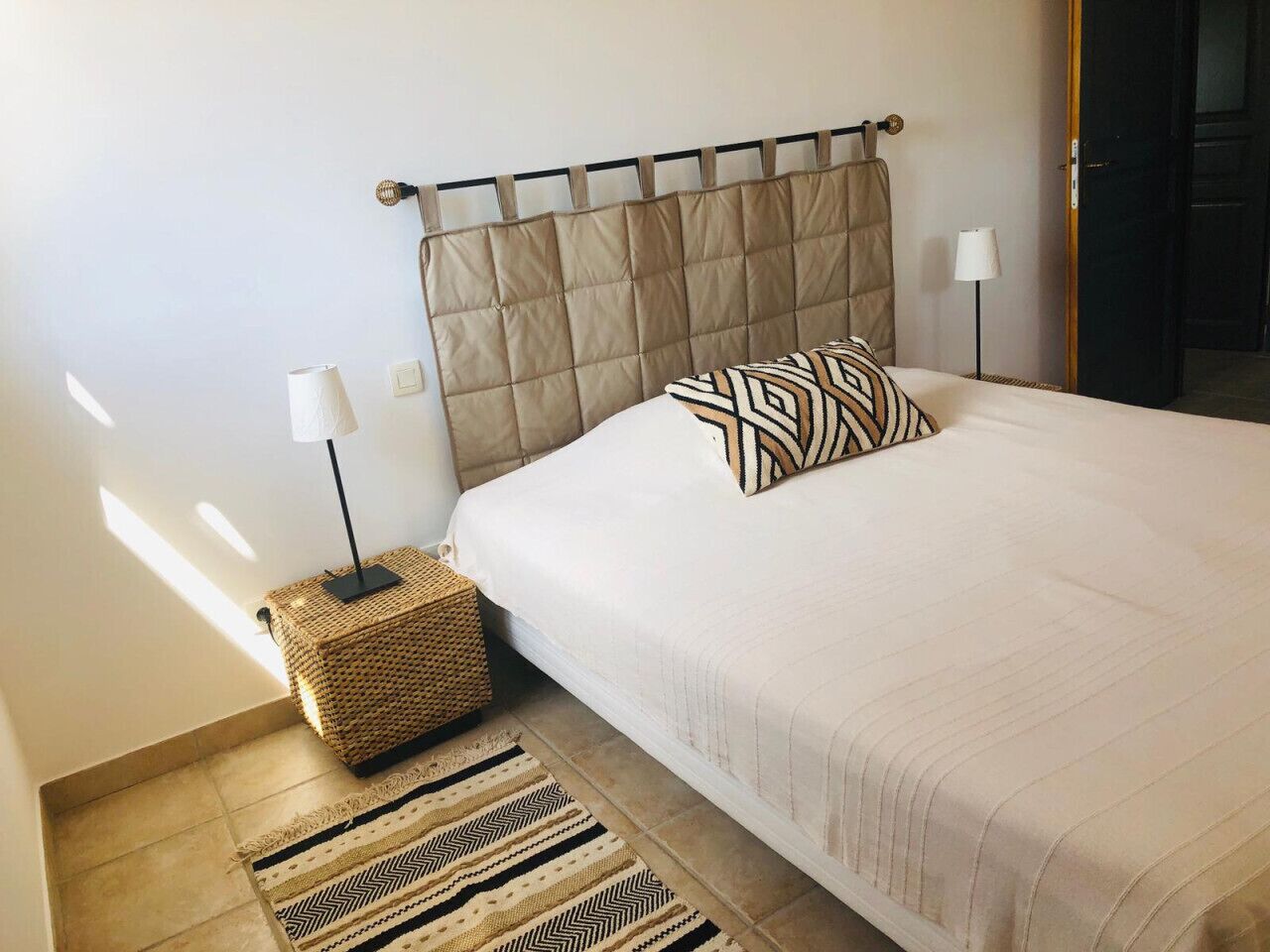 A neatly arranged bedroom with a bed, bedside lamps, a woven nightstand, and a patterned rug.