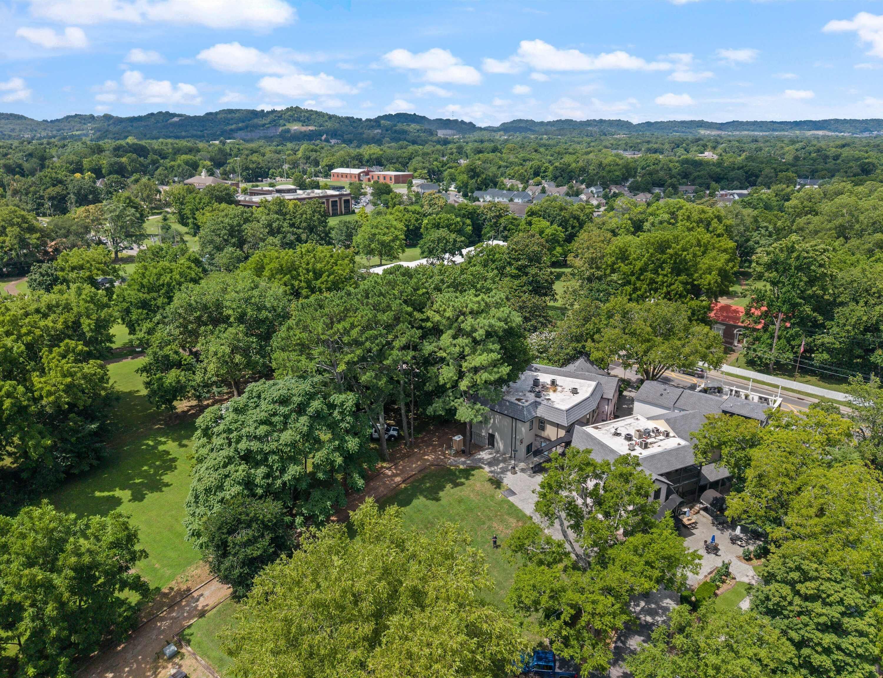 An aerial view of a residential area with houses, a large building with a flat roof, and surrounding greenery.