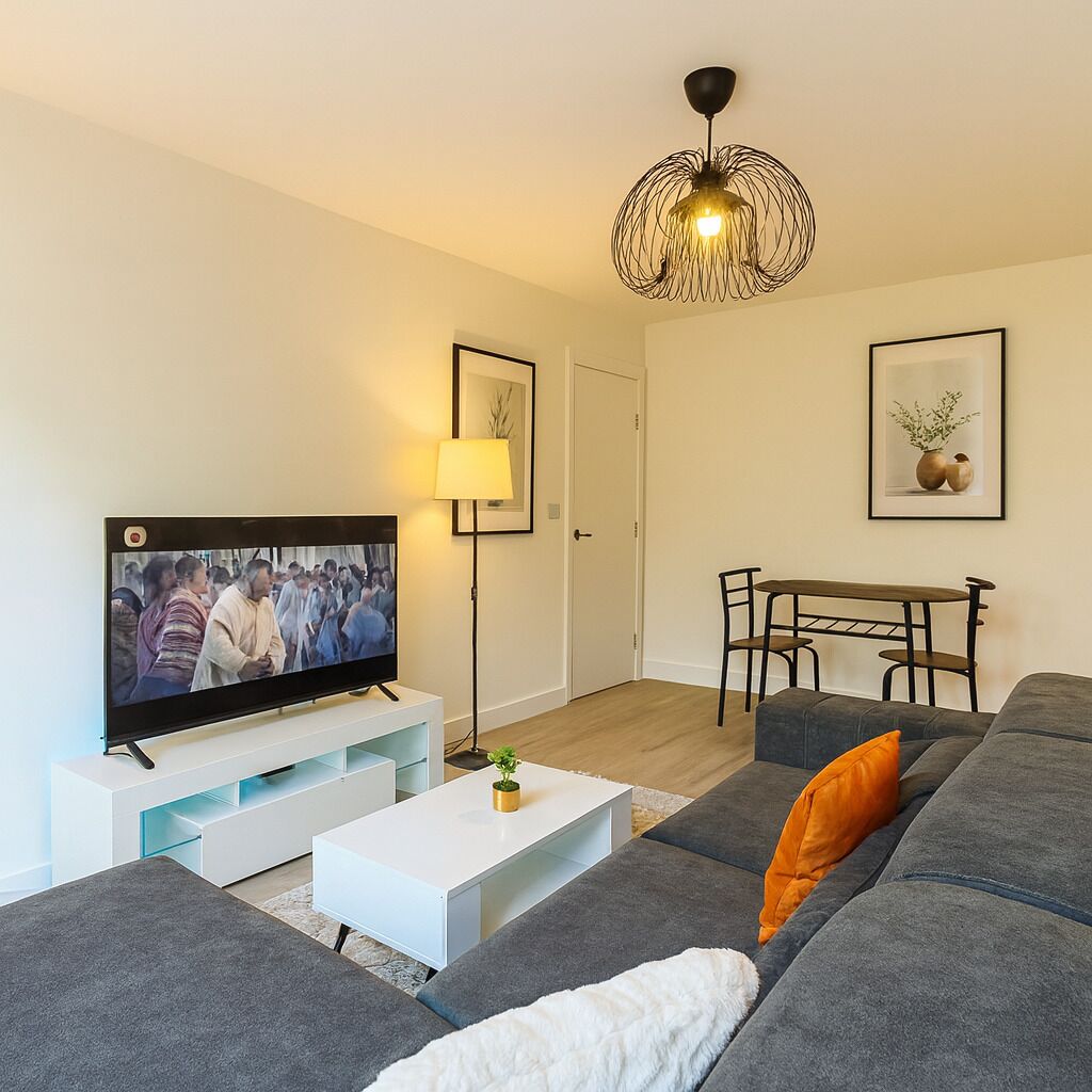 Una sala de estar moderna con un televisor de pantalla plana, una mesa de centro blanca, un sofá gris, una pequeña mesa con sillas y una lámpara colgante negra de diseño.