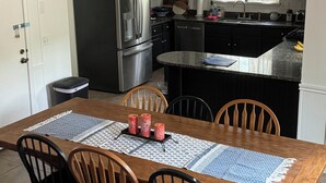 A dining area with a wooden table, four chairs, a chandelier, a refrigerator, a ceiling fan, and a kitchen with white cabinets and a black countertop. - Brookdale Blue sleeps 8, nestled in lakeside community in Highland Village, TX (Highland Village)