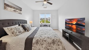 A bedroom with a bed, a ceiling fan, a TV, and a window with a view of houses.