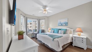 A bedroom with a bed, bedside tables, a ceiling fan, a TV mounted on the wall, and a window with curtains.