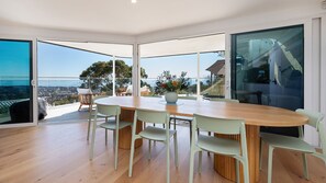 Ein moderner Essbereich mit einem Holztisch und hellgrünen Stühlen, große Glasüren führen zu einem Balkon mit Blick auf Stadt und Meer.