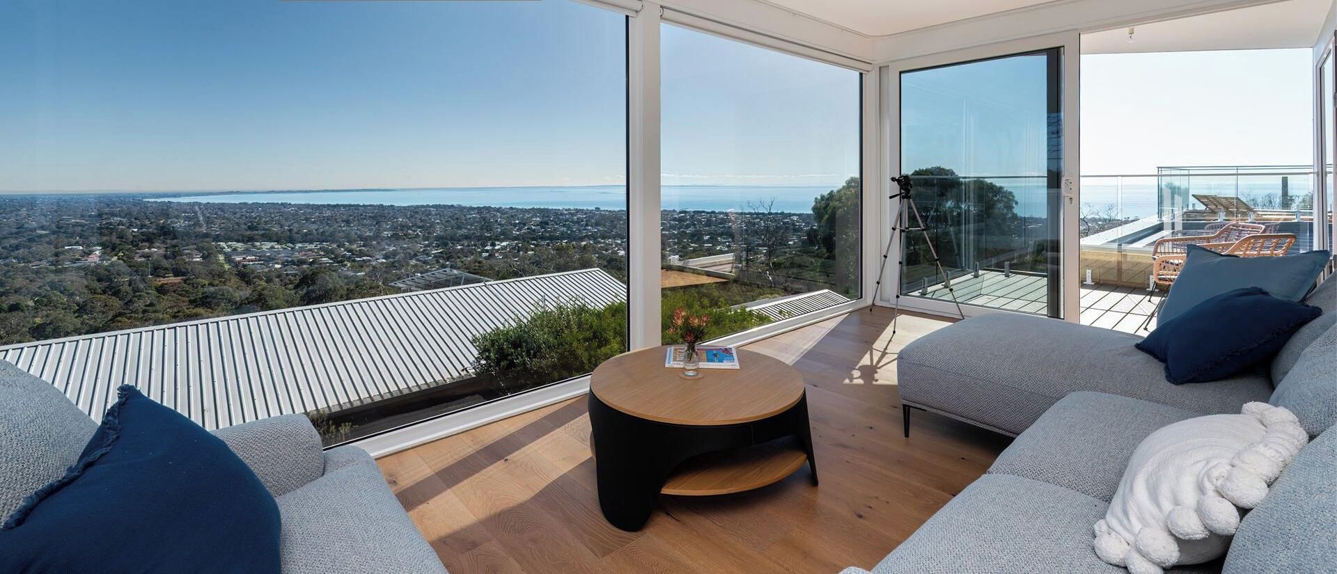 Ein modernes Wohnzimmer mit einem großen Glastür, einem runden Holztisch, einem grauen Sofa und Blick auf Stadt und Wasser.