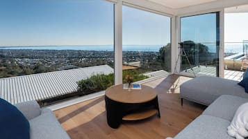 Ein modernes Wohnzimmer mit einem großen Glastür, einem runden Holztisch, einem grauen Sofa und Blick auf Stadt und Wasser.