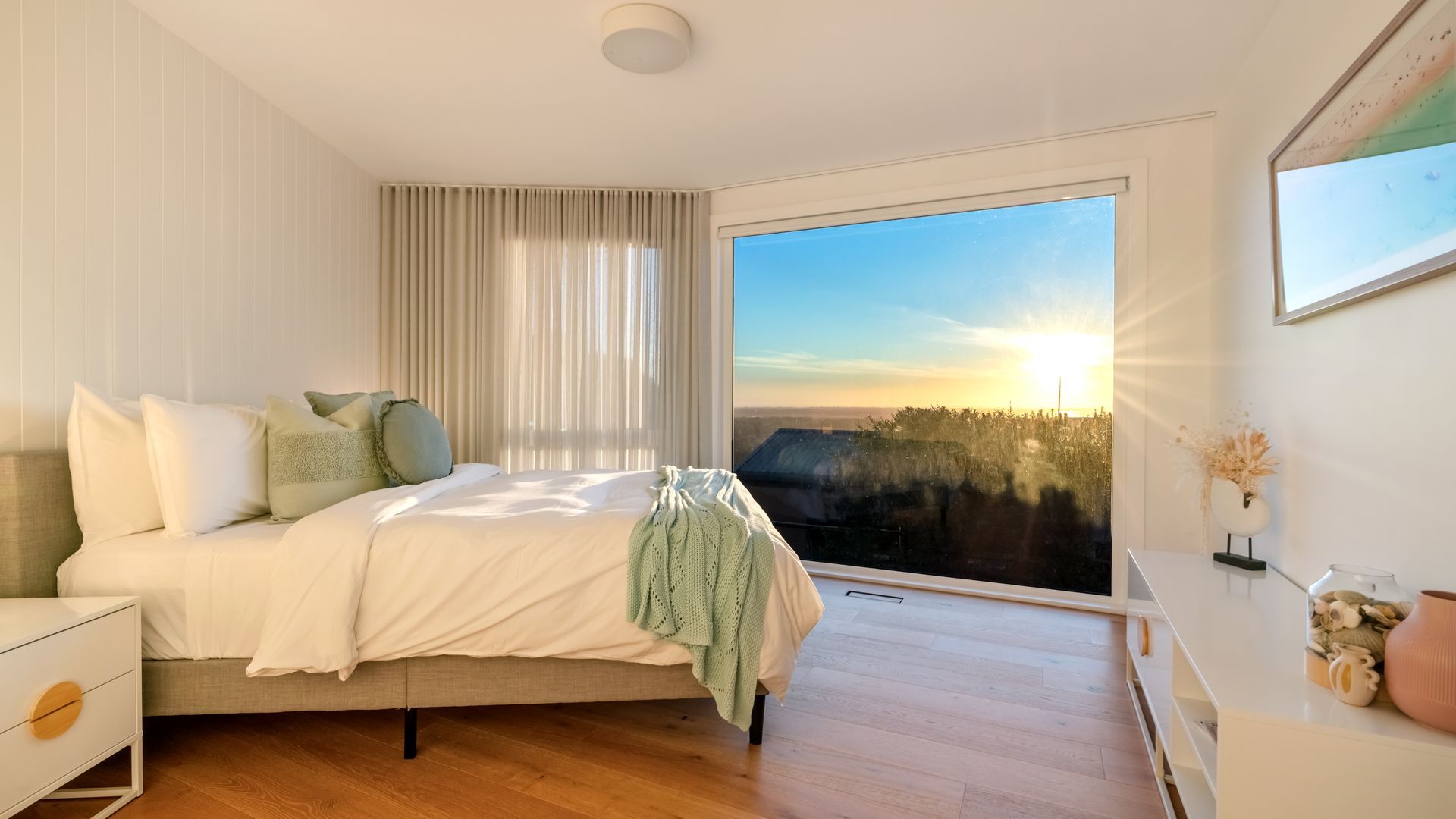 Ein Schlafzimmer mit einem Bett, einem Nachttisch, einer Kommode und einem großen Fenster mit Blick auf den Sonnenuntergang.