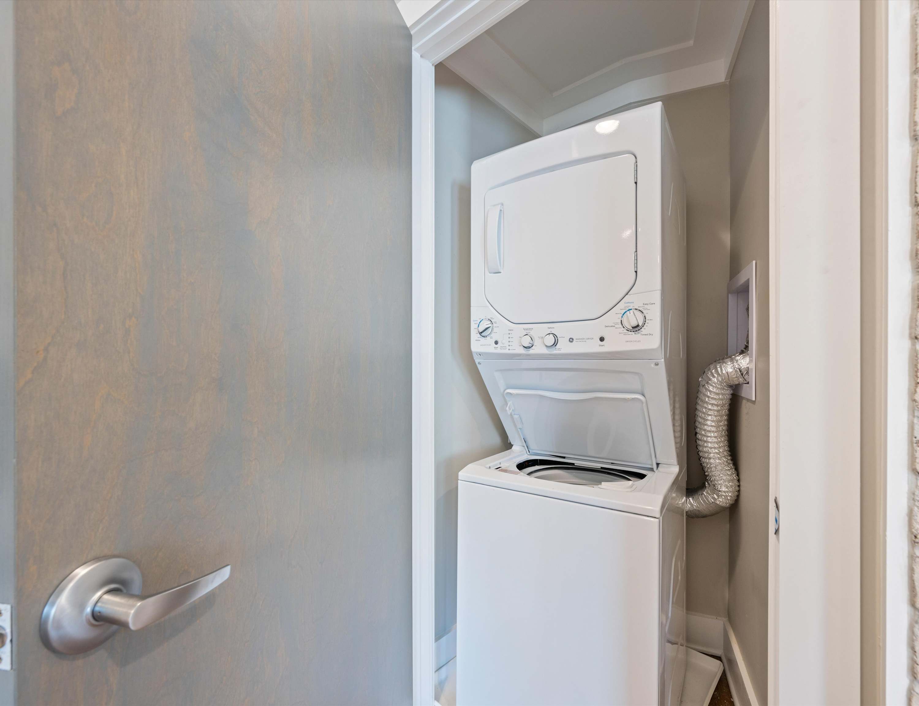 Une petite buanderie équipée d’un lave-linge et d’un sèche-linge intégrés, d’une porte avec une poignée argentée et d’un tuyau d’évacuation fixé au sèche-linge.