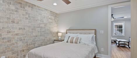 A bedroom with a brick wall, a wooden floor, a bed with a white bedspread, a nightstand with a lamp, and a ceiling fan.