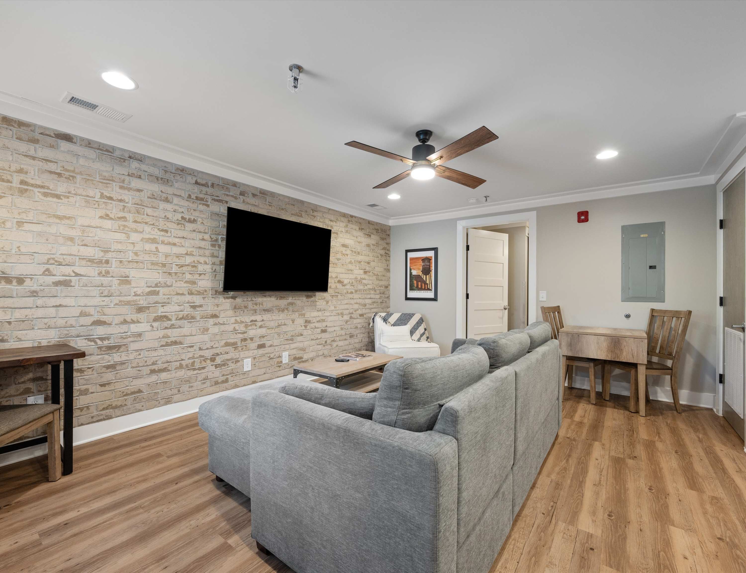 A living room with a brick wall, a flat-screen TV, a ceiling fan, a gray sofa, a wooden coffee table, and a dining area with a chair and table.