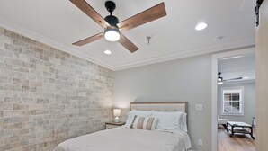 A bedroom with a brick wall, a wooden floor, a bed with a white bedspread, a nightstand with a lamp, and a ceiling fan. - Carters Court Cozy Hideaway (Franklin)