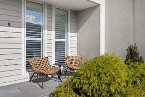 Un balcon avec deux fauteuils en osier et une petite table, un immeuble portant le numéro « 9 », et une haie au premier plan.