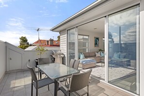 Een modern terras buiten met een glazen tafel en rieten stoelen, een barbecue en een schuifpui die leidt naar een leefruimte binnen.