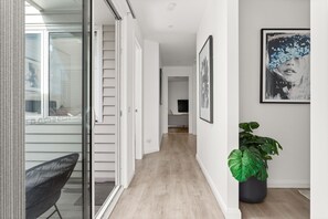 Un couloir avec une porte-fenêtre coulissante donnant sur un balcon, une plante en pot et des œuvres d’art encadrées accrochées au mur.