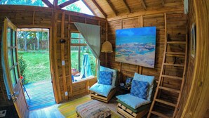 A cozy wooden cabin with a large window, a painting of a beach, and a bed with a colorful striped rug.