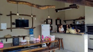 Una cocina rústica con techo de madera, un televisor montado en la pared y un refrigerador. Hay taburetes y una mesa con varios objetos encima.