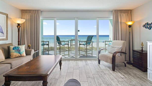 A room with a wooden coffee table, a sofa with a decorative pillow, a chair, and a view of the beach through large glass doors. - Beachfront Condo Cabana Club 202 Beautiful Water Views& Private Beach Access (Destin)