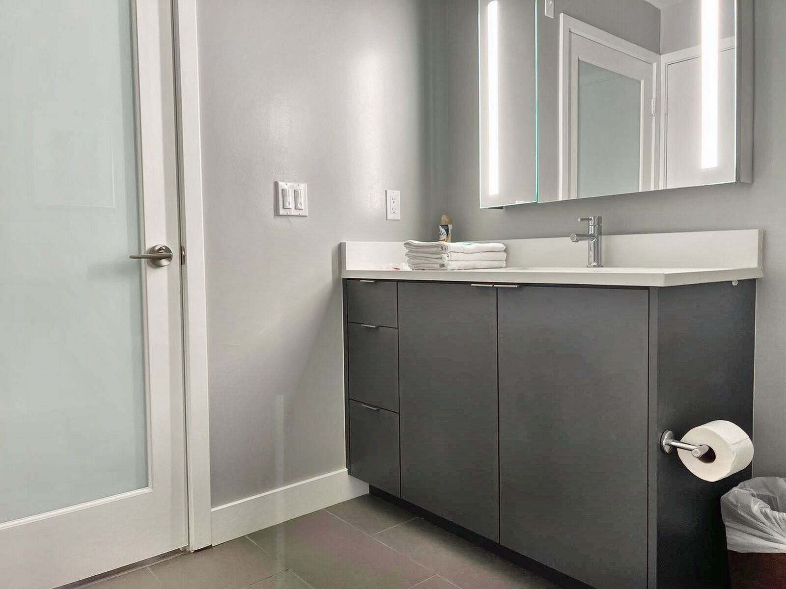 Un baño moderno con pared gris, puerta blanca, mueble gris oscuro, encimera blanca, espejo y dispensador de papel higiénico.