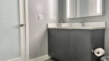 Un baño moderno con pared gris, puerta blanca, mueble gris oscuro, encimera blanca, espejo y dispensador de papel higiénico.