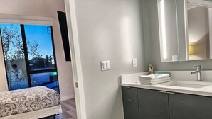 A modern bathroom with a large mirror, a sink, and a toilet. There is a bed with a patterned bedspread in the adjacent room. - Cozy Modern Studio on Spaulding Ave – Complimentary WiFi & Parking (Los Angeles)