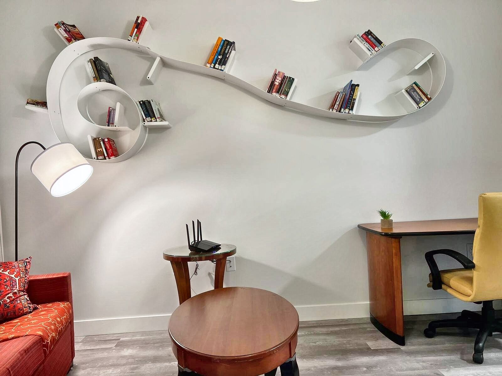 Une pièce moderne avec une bibliothèque aux étagères incurvées, une table ronde en bois, un canapé rouge, un bureau avec une chaise et un lampadaire.