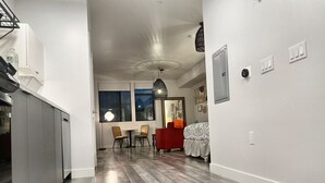 A modern kitchen with grey cabinets, a dining area with a red chair, and a bedroom with a patterned bedspread. - Cozy Modern Studio on Spaulding Ave – Complimentary WiFi & Parking (Los Angeles)