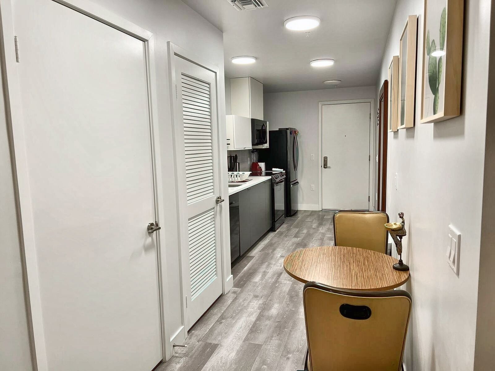 A narrow hallway with a kitchen area on the right, featuring a refrigerator, stove, and cabinets. There are two chairs and a small table in the hallway.