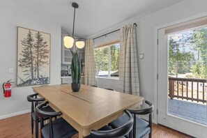 Un espace repas avec une table en bois, des chaises noires, une plante en pot, des luminaires suspendus, un extincteur et une vue sur l’extérieur par une fenêtre.