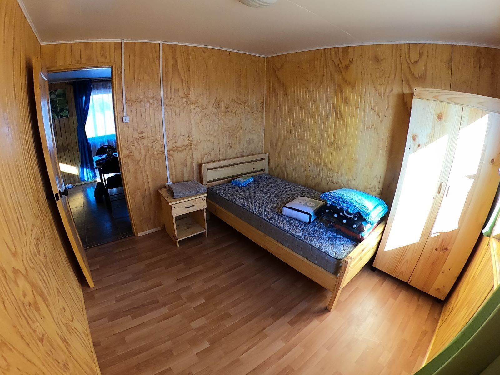 Ein Zimmer mit Holzwänden, einem Bett, einem Nachttisch, einem Kleiderschrank und einer Tür zu einem anderen Raum.
