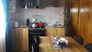 Een kleine keuken met houten kastjes, een fornuis, een spoelbak en een eettafel met stoelen.
